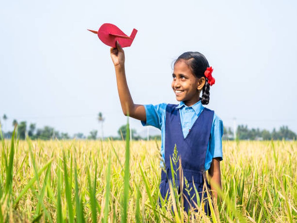 Happy village girl kid with school uniform playing using toy cardboard airplane at paddy field - concept of childhood dream, aspirations and freedom